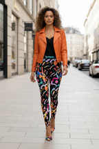 A woman walking down a city street wearing a bright orange leather jacket, a black top, and colorful abstract-print leggings with orange pointed-toe heels.
