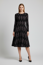 A woman stands against a light gray studio background wearing a long-sleeve black midi dress with an allover tonal floral pattern and black pointed heels.