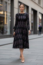 A woman stands on a city sidewalk wearing a long-sleeve midi dress with a black base and an allover pink graphic print. The dress has a round neckline and a flared skirt.