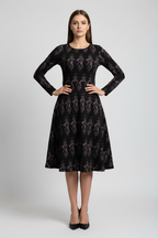 A woman stands against a light gray studio background wearing a long-sleeve black dress with an allover tonal floral pattern and black pointed-toe heels.