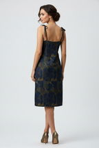A woman modeling a sleeveless knee-length dress from the back, featuring a dark navy blue and gold abstract patterned fabric, thin shoulder ties, and metallic gold heels against a light gray studio background.