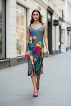 A woman walks down a city street wearing a sleeveless midi dress with thin straps and a colorful abstract floral print on a muted blue-gray background, paired with bright pink heels.