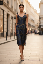 A woman walks down a city street wearing a fitted midi dress with thin straps and a dark blue and gold abstract pattern, paired with clear high-heel sandals.