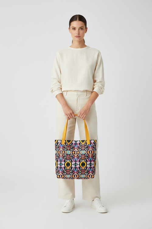 A model stands against a light gray studio background wearing a cream sweatshirt, cream pants, and white sneakers, while holding a colorful geometric patterned tote bag with yellow handles.