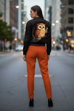 A woman stands on a city street facing away from the camera, wearing a black bomber-style jacket with a large owl graphic on the back and bright orange pants.