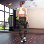 Woman standing, wearing black leggings with an intricate golden pattern and a white sports bra.