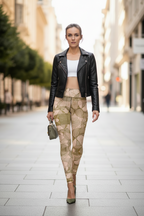 A woman walking down a city street wearing a black leather moto jacket, a white crop top, beige and olive green patterned leggings, and green heels, holding a small green handbag.