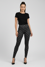 A woman modeling black patterned leggings with a black short-sleeve top and black heels, standing against a light gray studio background.