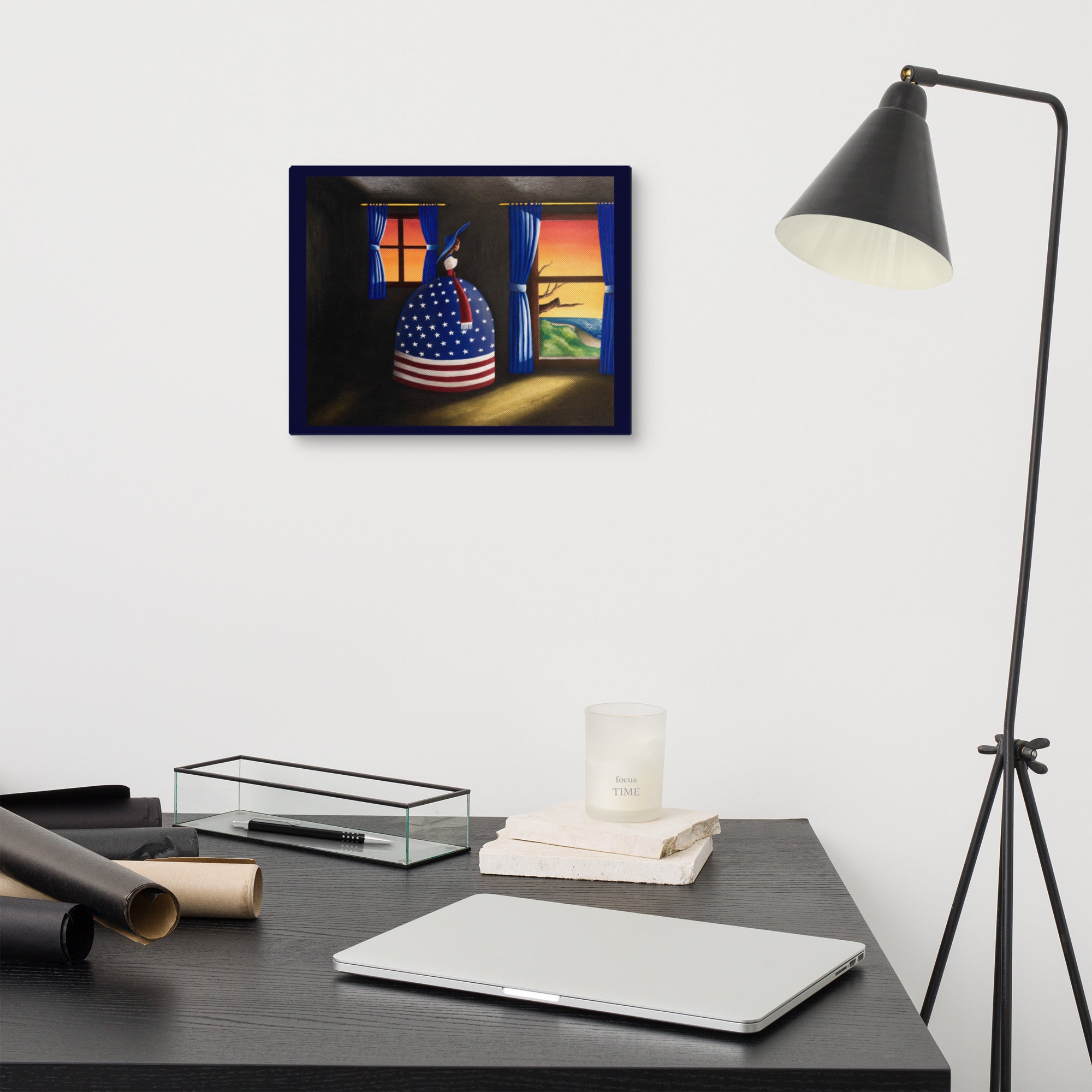 A framed art print depicting a woman in a patriotic American dress under a window at sunset, displayed on a white wall above a dark desk with a laptop, candle, and lamp.
