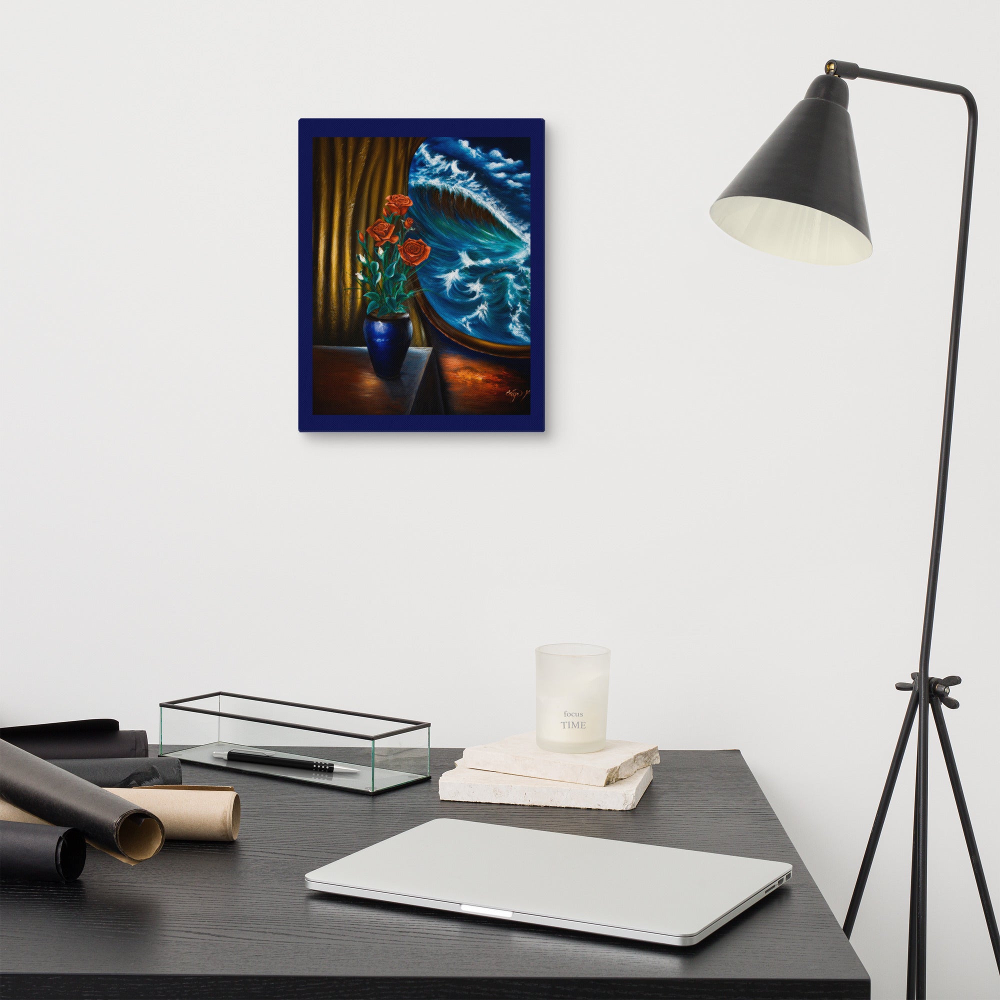 A framed art print depicting a vibrant still life with red roses, a blue vase, and a blue-green wave, displayed on a white wall above a dark desk with a laptop, lamp, and other office accessories.