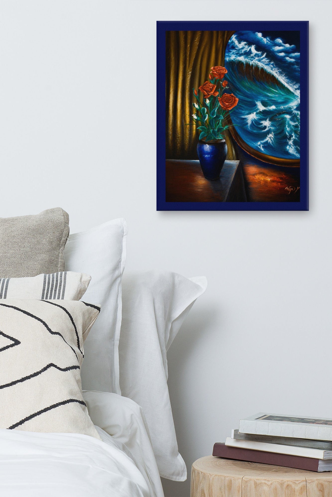 A framed art print depicting a blue ocean wave crashing against a shore, with a blue vase of red flowers in the foreground, displayed on a light gray wall above a bed with white linens and a small wooden side table with books.