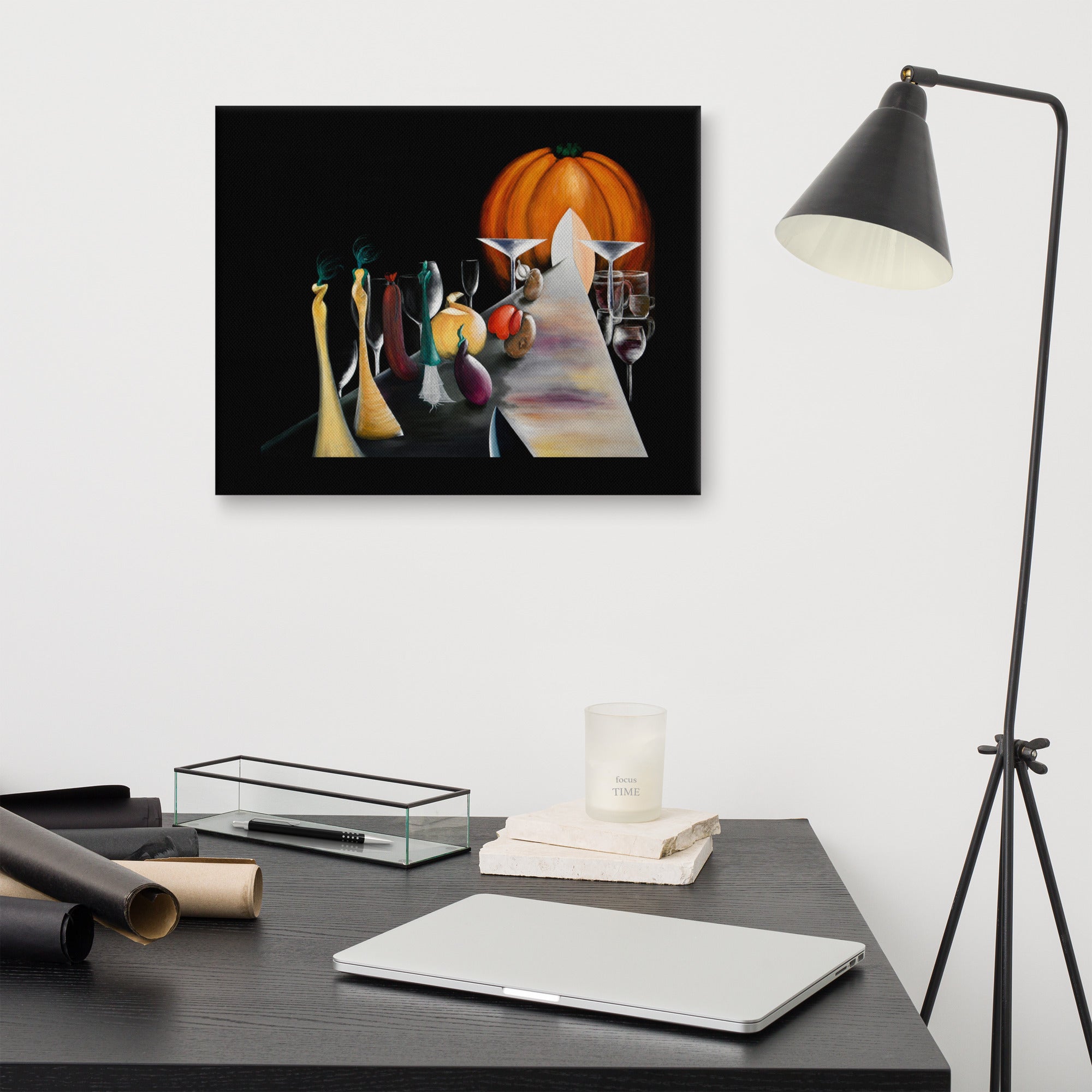 A framed wall art print depicting a stylized still life with a large orange pumpkin and several tall yellow glasses, displayed above a dark desk in a modern room with a black floor lamp, laptop, and rolled papers.
