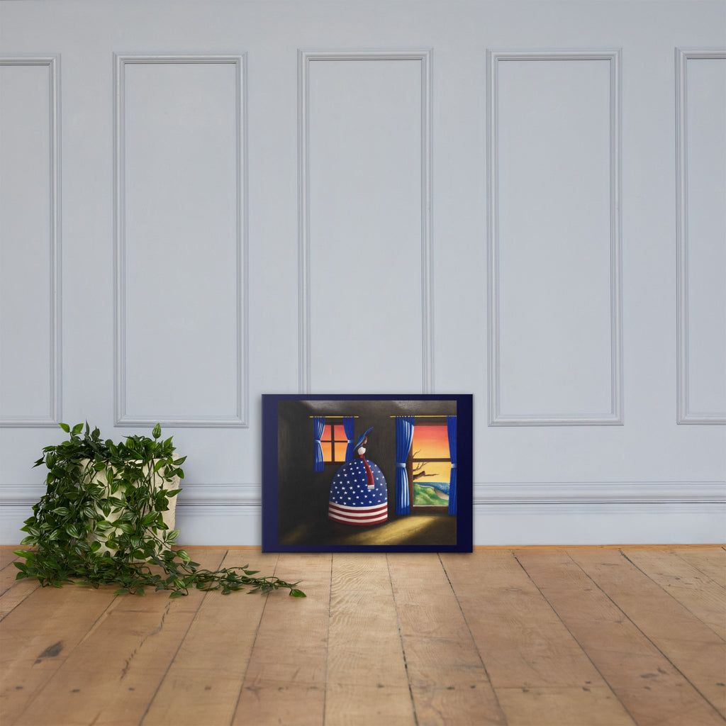 A framed art print leaning against a white paneled wall on a light wood floor, next to a potted trailing green plant. The artwork depicts a whimsical scene of a donkey wearing an American flag pattern and looking out of a stable at sunset.