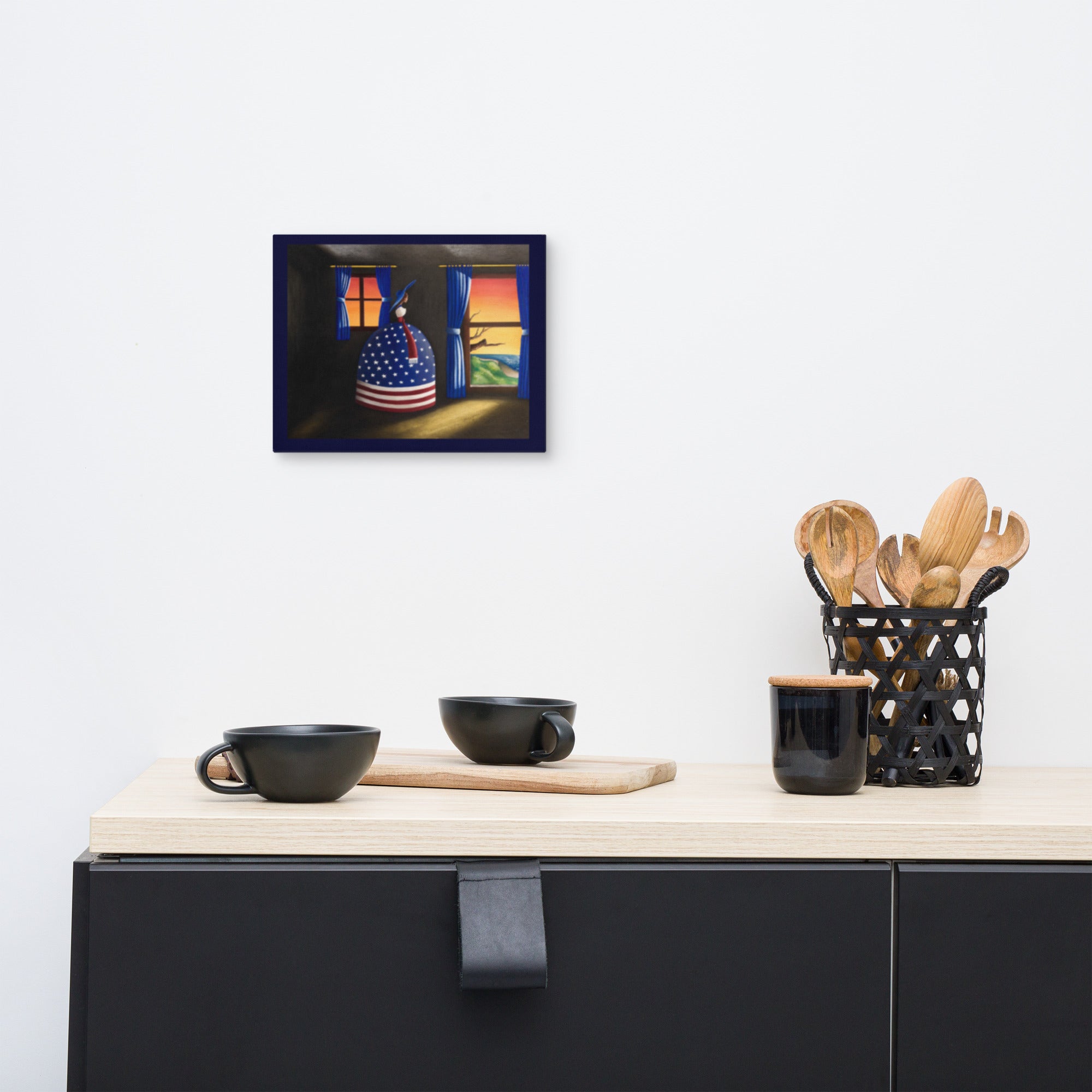 A framed wall art print depicting a colorful still life with an American flag patterned pot and warm glowing windows, displayed on a white wall above a kitchen counter with black mugs and utensils.