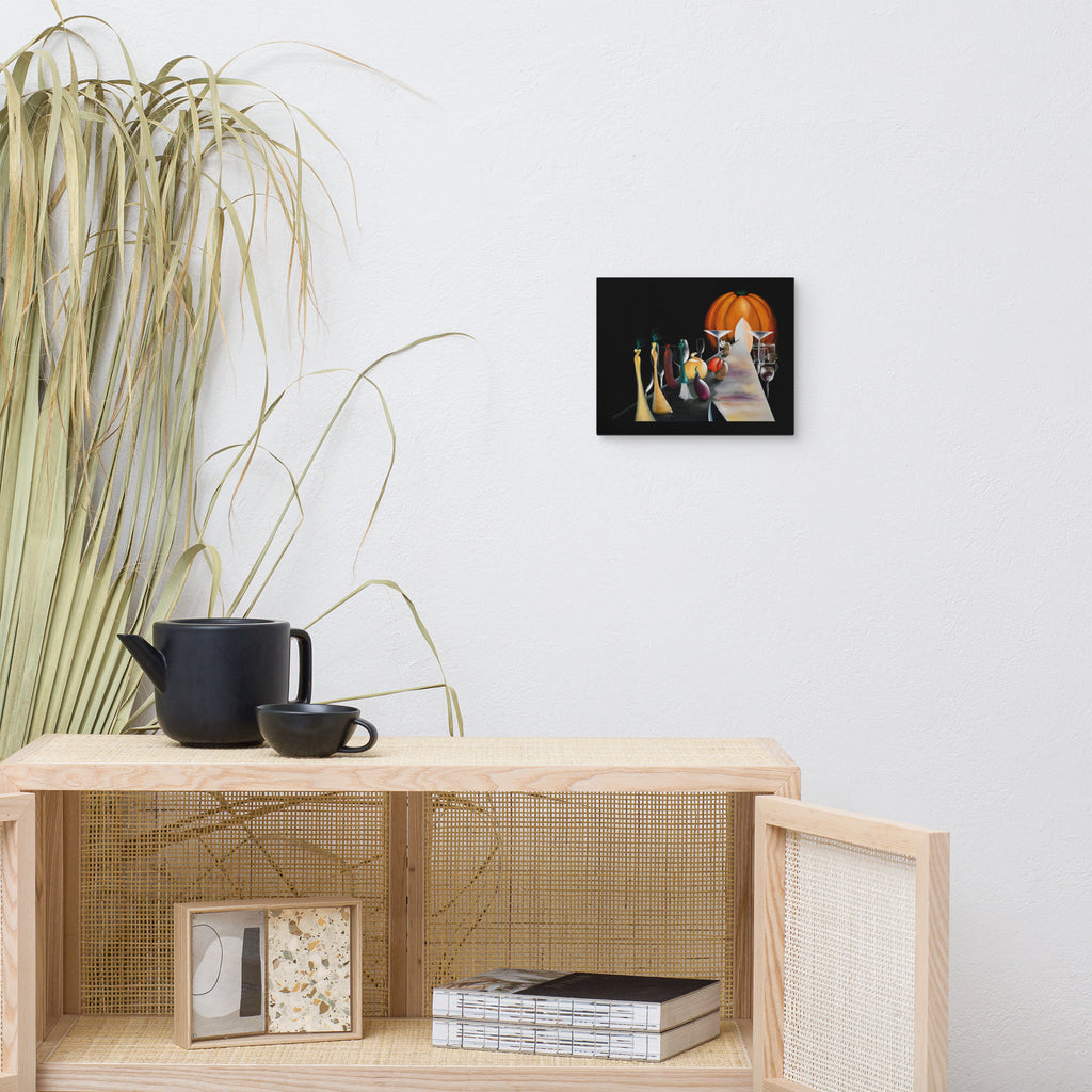 A small framed art print depicting a stylized still life with a pumpkin and candles, mounted on a white wall above a light wood shelf with a black teapot and dried grass decor.