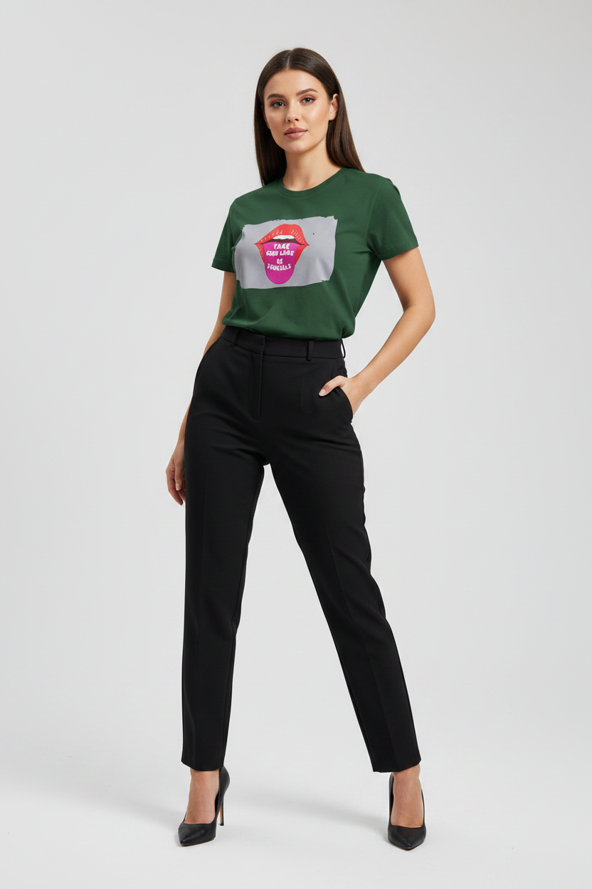 A woman modeling a dark green short-sleeve graphic T-shirt with a pink and gray lip design, paired with black trousers and black high heels, against a light gray studio background.