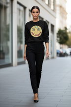 A woman walking down a city street wearing a black crewneck sweatshirt with a large circular graphic on the chest, paired with black pants and black heels.