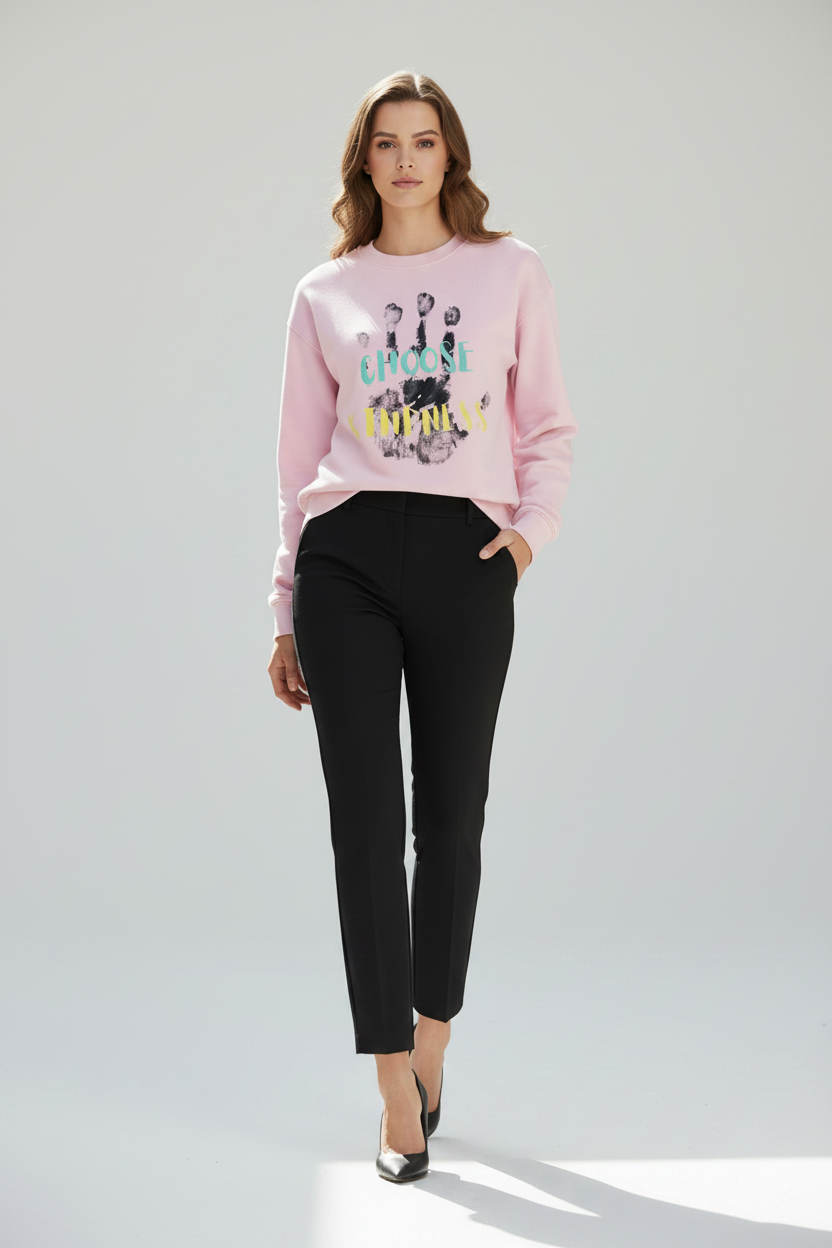 A woman modeling a light pink crewneck sweatshirt with a colorful graphic print on the front, paired with black cropped trousers and black pointed-toe heels, against a light gray studio background.