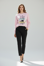 A woman modeling a light pink crewneck sweatshirt with a colorful graphic print on the front, paired with black cropped trousers and black pointed-toe heels, against a light gray studio background.