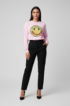 A model stands against a light gray studio background wearing a light pink crewneck sweatshirt with a large yellow and green graphic on the front, paired with black trousers and black heels.