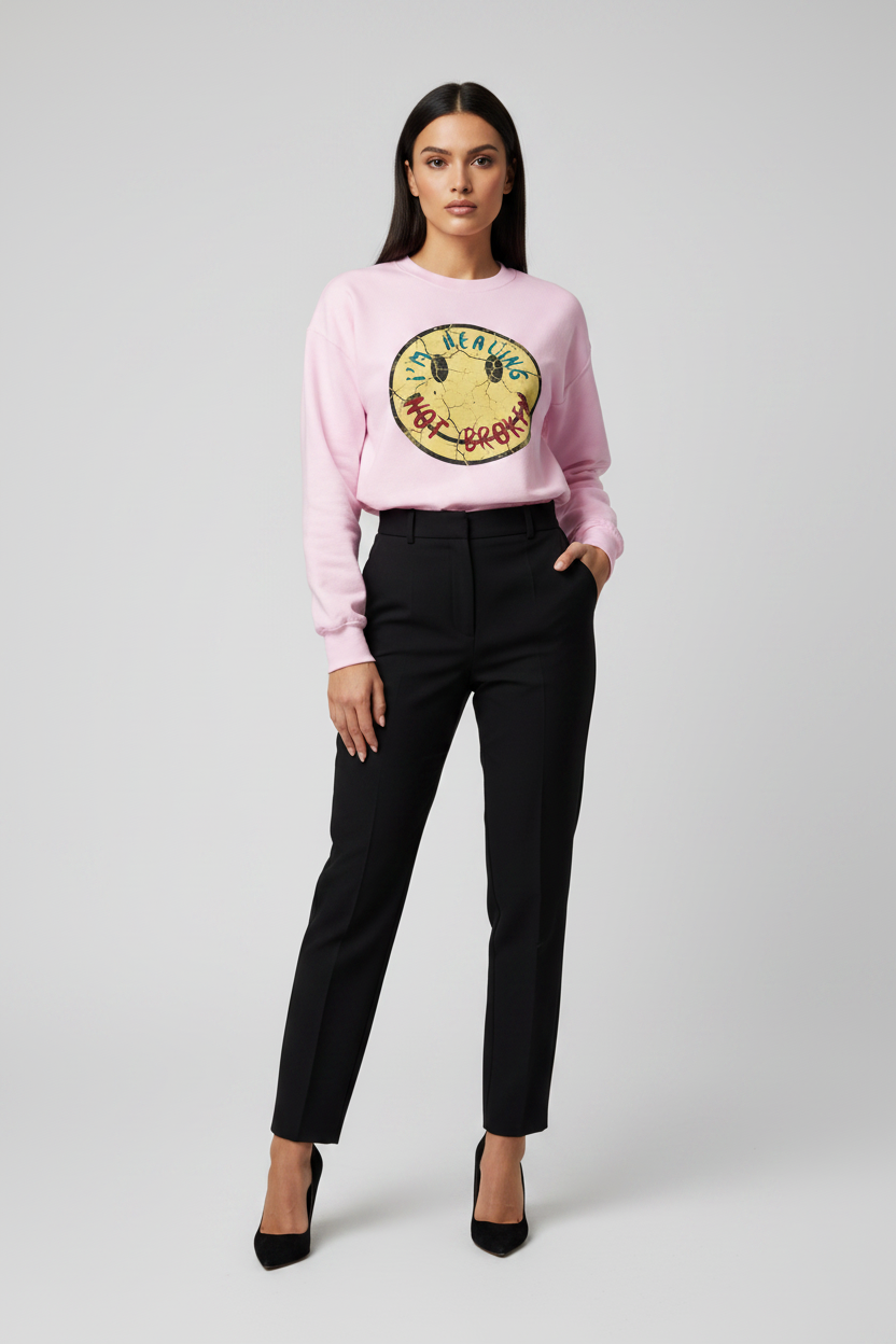 A model stands against a light gray studio background wearing a light pink crewneck sweatshirt with a large yellow and green graphic on the front, paired with black trousers and black heels.