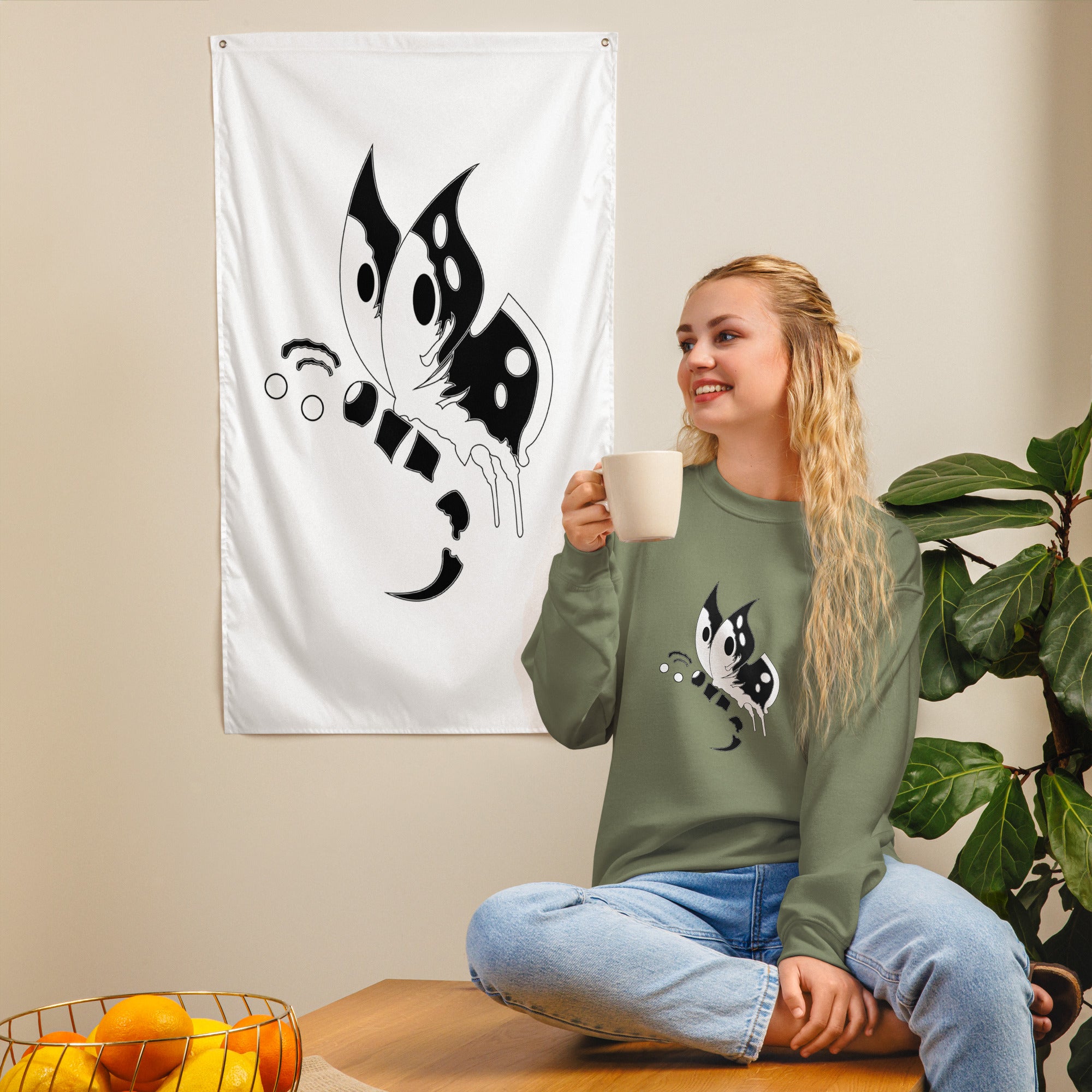 A woman sitting and smiling, wearing a green long-sleeve sweatshirt with a black graphic print of a butterfly on the front, holding a coffee cup, with a white banner with a butterfly graphic in the background.