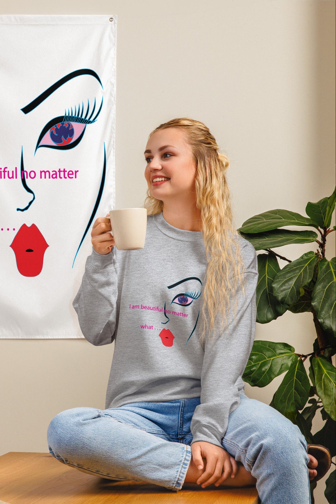 A woman sitting and drinking from a mug, looking at a white abstract art sweatshirt with a text and face design, displayed on a wall.