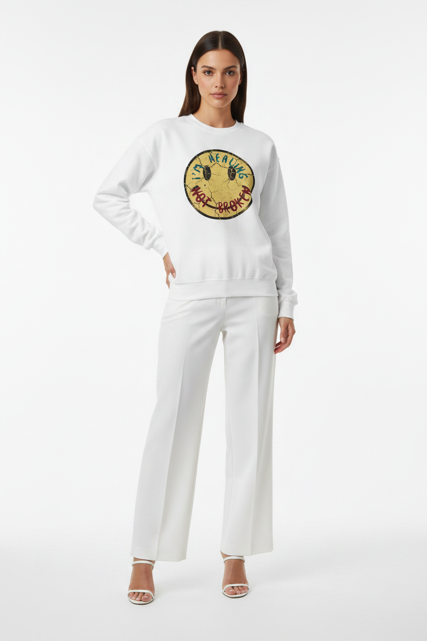 A model wearing a white crewneck sweatshirt with a circular embroidered smiley face graphic on the chest, paired with matching white wide-leg pants and white heeled sandals, against a plain white studio background.