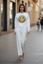 A woman walking down a city street wearing a white crewneck sweatshirt with a large circular graphic on the chest and matching white wide-leg pants.