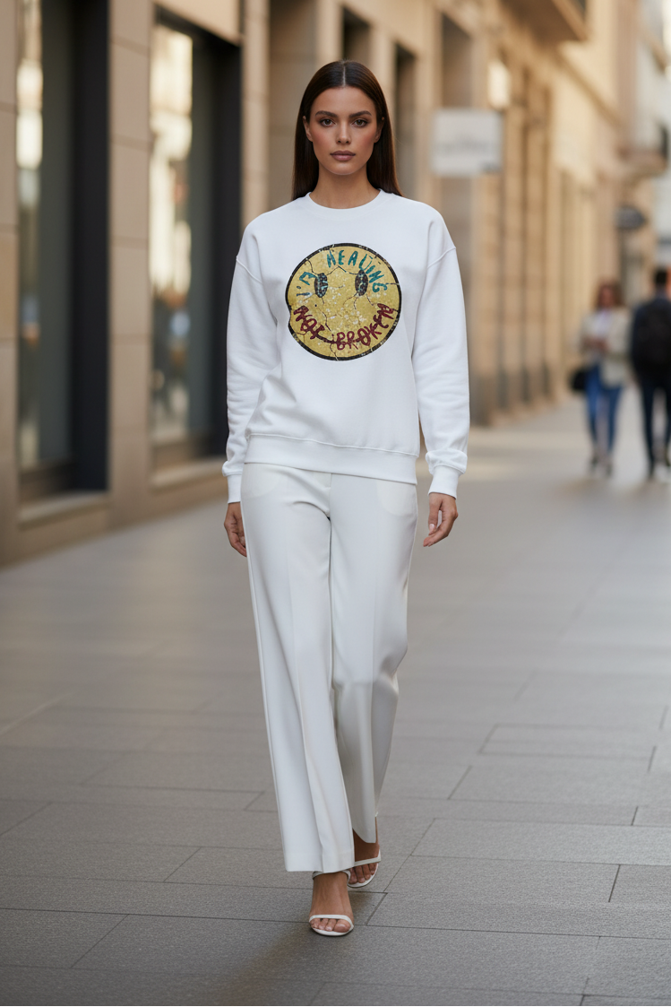A woman walking down a city street wearing a white crewneck sweatshirt with a large circular graphic on the chest and matching white wide-leg pants.