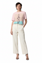 A model stands against a plain white studio background wearing a short-sleeve graphic T-shirt with a pink, beige, and turquoise color-block design, paired with wide-leg cream trousers and black heeled sandals.