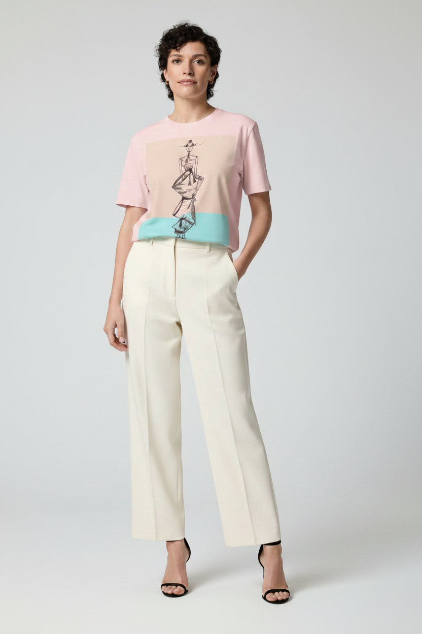 A model stands against a light gray studio background wearing a pastel graphic T-shirt with pink, beige, and turquoise color blocks, paired with wide-leg cream trousers and black heeled sandals.
