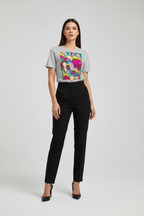 A woman modeling a light gray short-sleeve t-shirt with a colorful geometric graphic print on the front, paired with black trousers and black heels, against a light gray studio background.