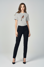A woman modeling a light gray short-sleeve t-shirt with a small bird graphic on the left chest, paired with black pants and black heels, against a light gray studio background.