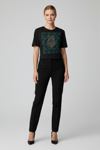 A woman modeling a black short-sleeve crewneck t-shirt with a dark teal and gold owl graphic on the front, paired with black trousers and black heels, against a light gray studio background.