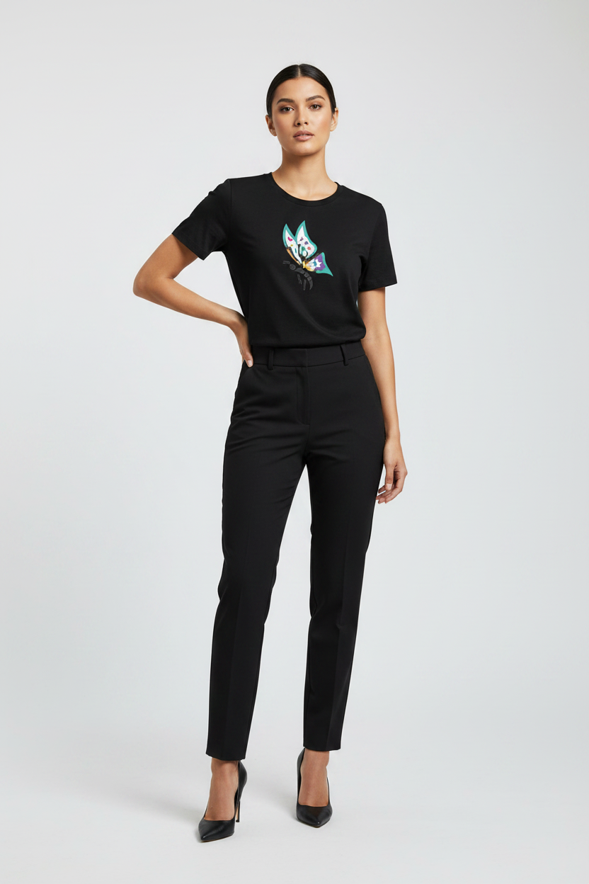 A woman modeling a black short-sleeve crewneck t-shirt with a colorful graphic on the chest, paired with black trousers and black heels, against a light gray studio background.