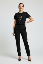 A woman modeling a black short-sleeve crewneck t-shirt with a colorful graphic print on the chest, paired with black trousers and black heels, against a light gray studio background.