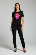 A woman modeling a black short-sleeve graphic T-shirt with a colorful lips design and the words 'SPEAK UP' in pink, paired with black trousers and black heels, against a light gray studio background.