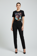 A woman modeling a black short-sleeve t-shirt with a colorful graphic print on the front, paired with black trousers and black heels, against a light gray studio background.