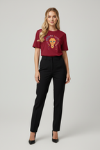 A woman modeling a short-sleeve crewneck t-shirt in deep burgundy with a colorful embroidered lion graphic on the chest, paired with black trousers and black heels, against a light gray studio background.