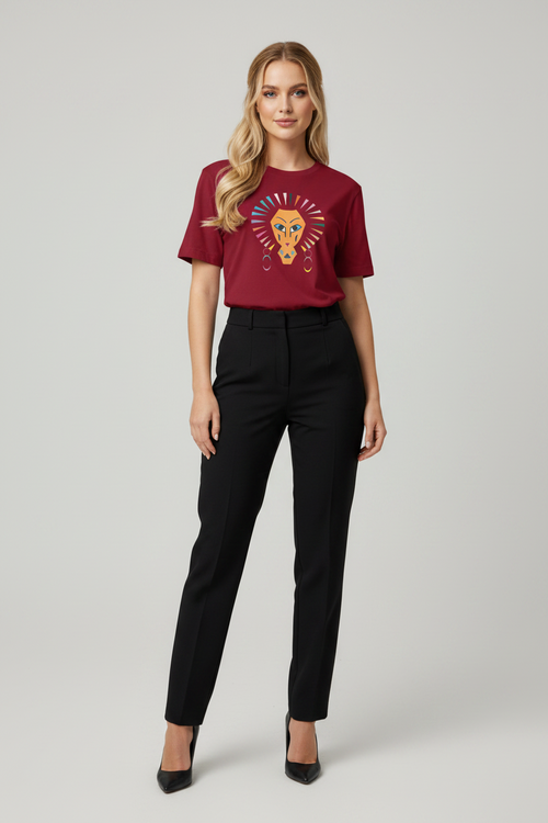 A woman modeling a short-sleeve crewneck t-shirt in deep burgundy with a colorful embroidered lion graphic on the chest, paired with black trousers and black heels, against a light gray studio background.