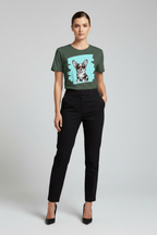 A model wearing a dark green short-sleeve t-shirt with a graphic print of a dog on a light blue background, paired with black trousers and black heels, against a light gray studio background.
