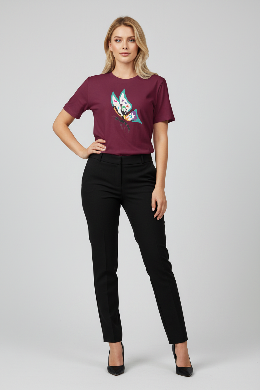 A woman modeling a short-sleeve crewneck t-shirt in deep burgundy with a colorful graphic print on the front, paired with black trousers and black heels, against a light gray studio background.