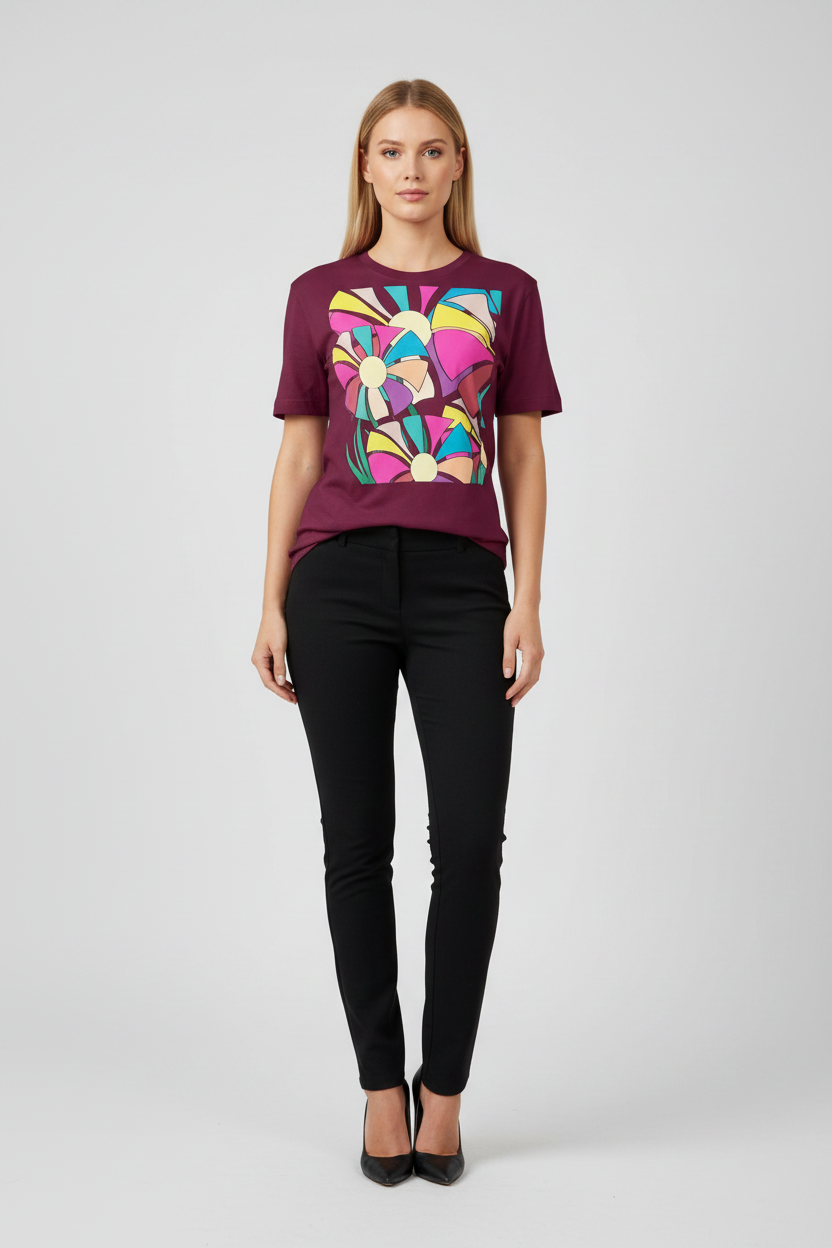A woman modeling a short-sleeve crewneck t-shirt in deep burgundy with a large, colorful abstract geometric graphic on the front, paired with black pants and black heels, against a light gray studio background.