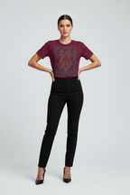 A woman modeling a short-sleeve crewneck t-shirt in deep burgundy with a red and blue geometric graphic print on the front, paired with black pants and black heels, against a light gray studio background.
