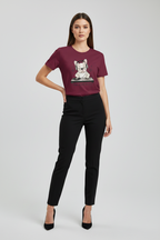 A woman modeling a short-sleeve crewneck t-shirt in burgundy with a cartoon dog graphic on the chest, paired with black pants and black heels, against a light gray studio background.