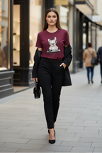 A woman walking on a city street wearing a maroon graphic t-shirt with a white cat illustration, black trousers, and a black blazer, holding a small black handbag.