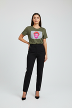 A woman modeling a short-sleeve olive green graphic t-shirt with a pink lips print and black high-waisted trousers, standing against a light gray studio background.