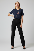A woman modeling a navy blue short-sleeve t-shirt with a colorful bird graphic on the chest, paired with black trousers and black heels, against a light gray studio background.