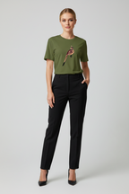 A woman modeling a short-sleeve olive green t-shirt with a bird graphic on the chest, paired with black trousers and black heels, against a light gray studio background.